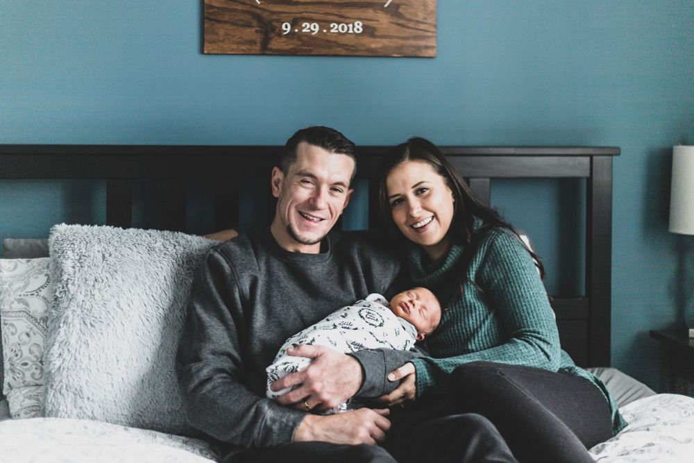 a shot of new parents sitting on their bed holding their newborn baby boy smiling and looking at the camera