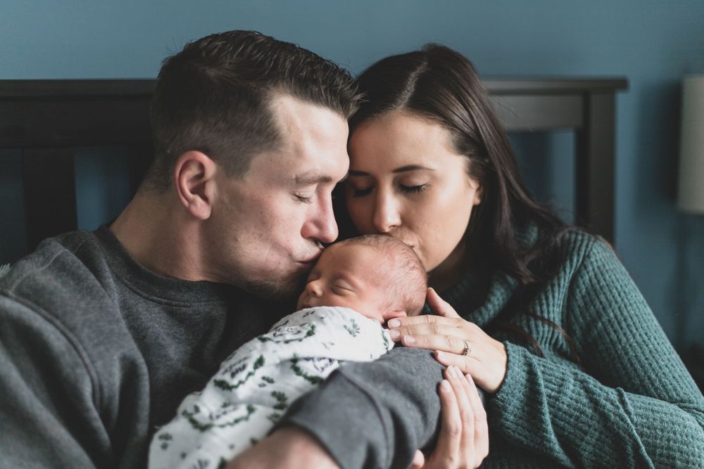 a shot of new parents sitting on their bed holding their newborn baby boy both kissing his head