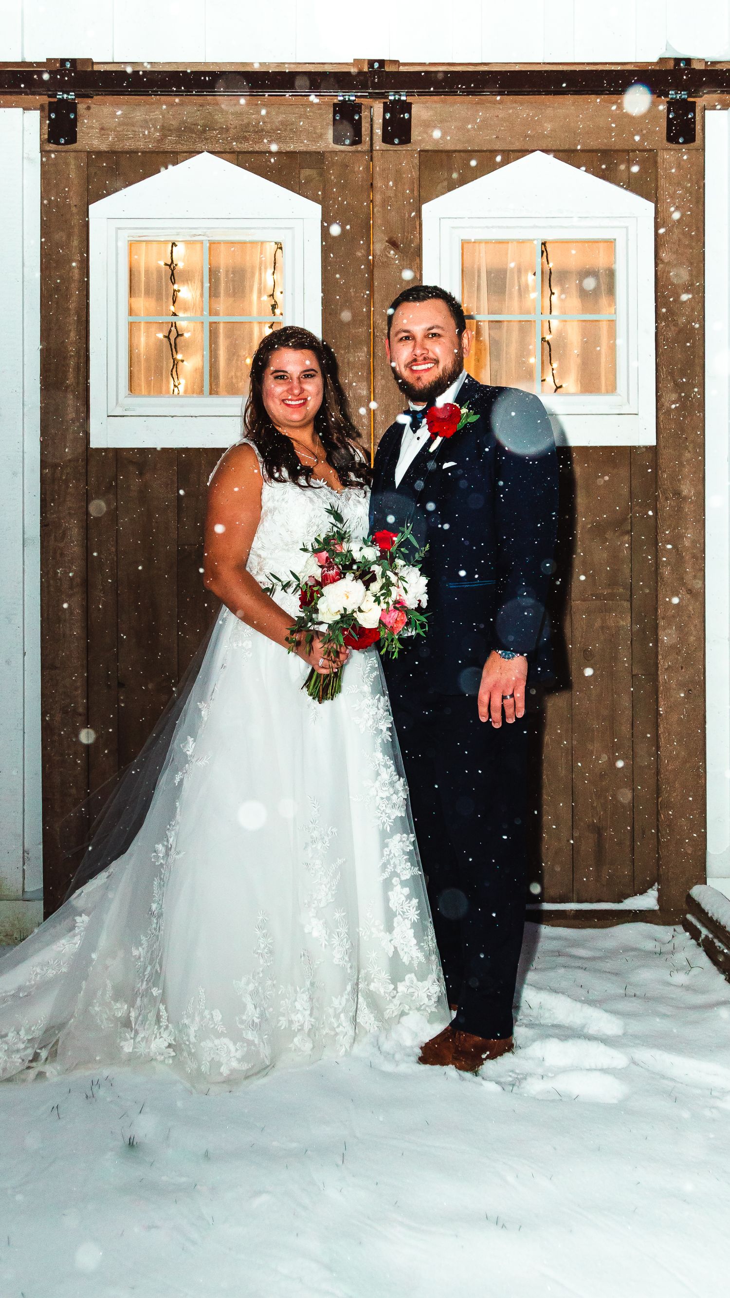 Winter wedding portrait of newlyweds standing in snow in front of rustic barn doors.