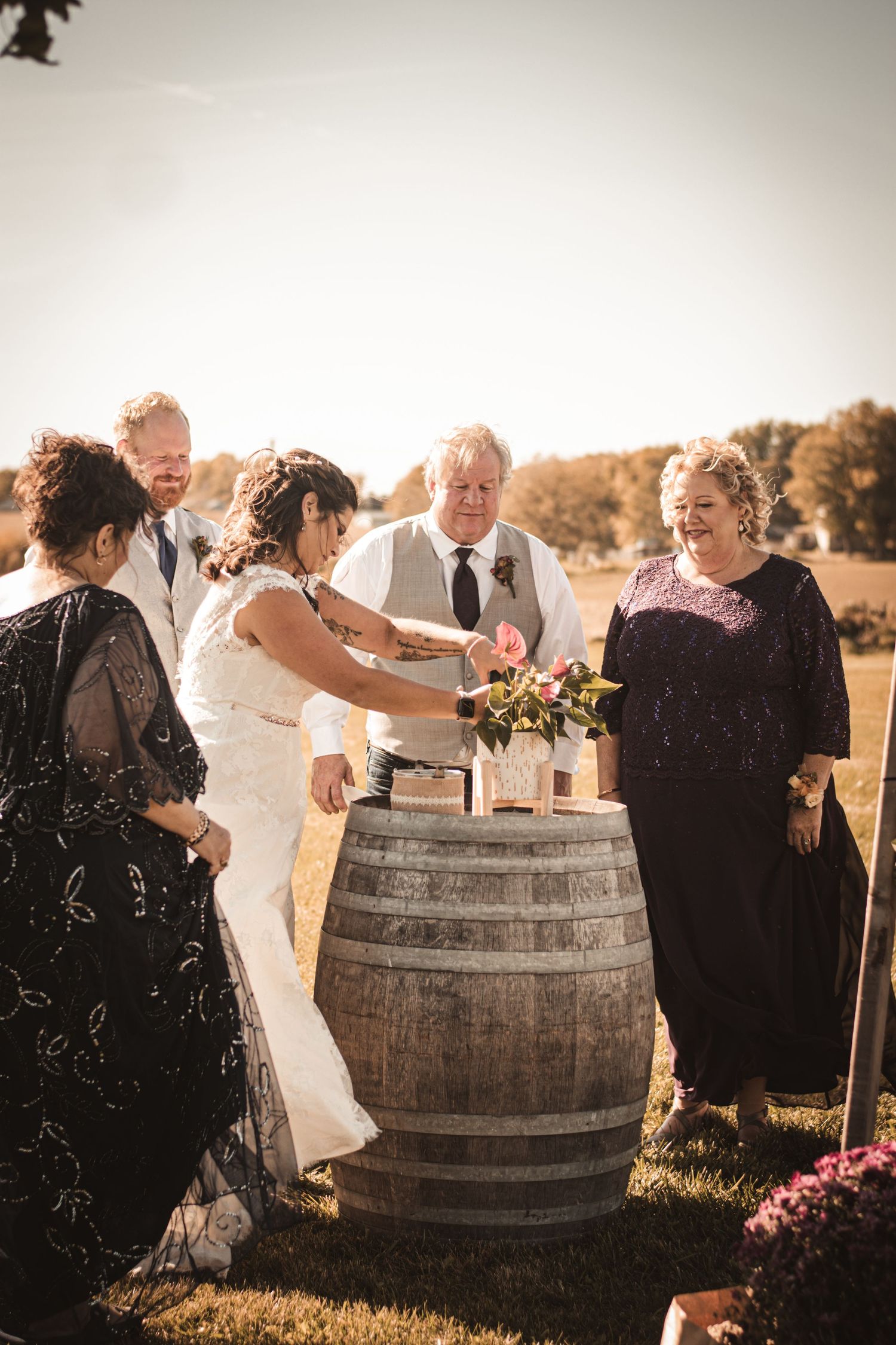 Wedding ceremony participants gather around rustic wine barrel outdoors with autumn florals.