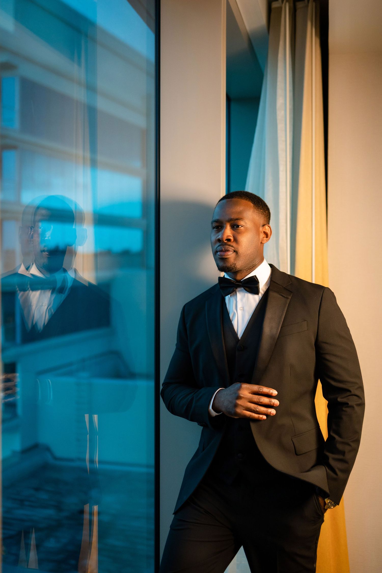 Groom in classic black tuxedo poses against window with dramatic blue lighting.