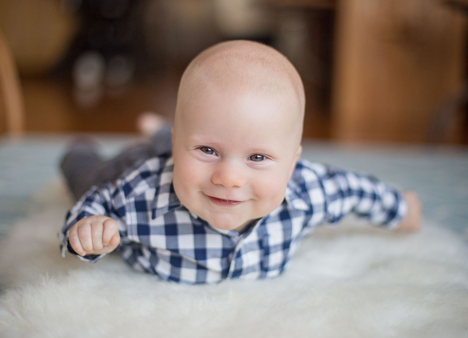 Baby wearing blue plaid shirt lying on white fluffy carpet smiles at camera.