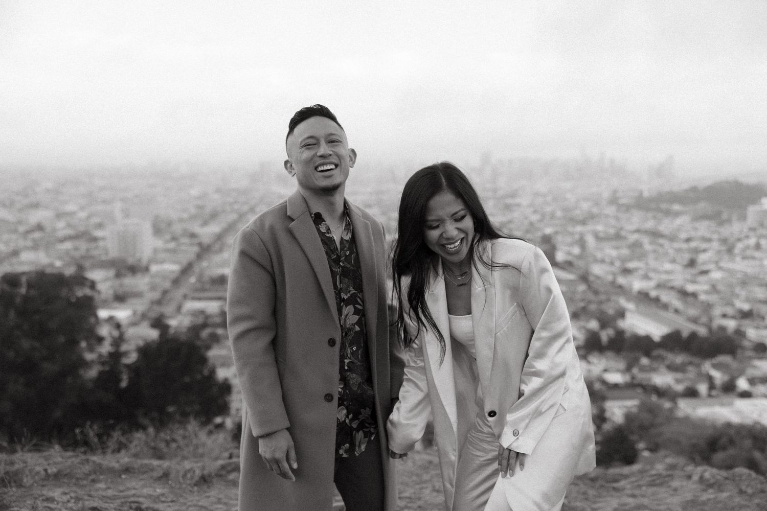 A candid black and white portrait captured on a hilltop overlooking a cityscape.