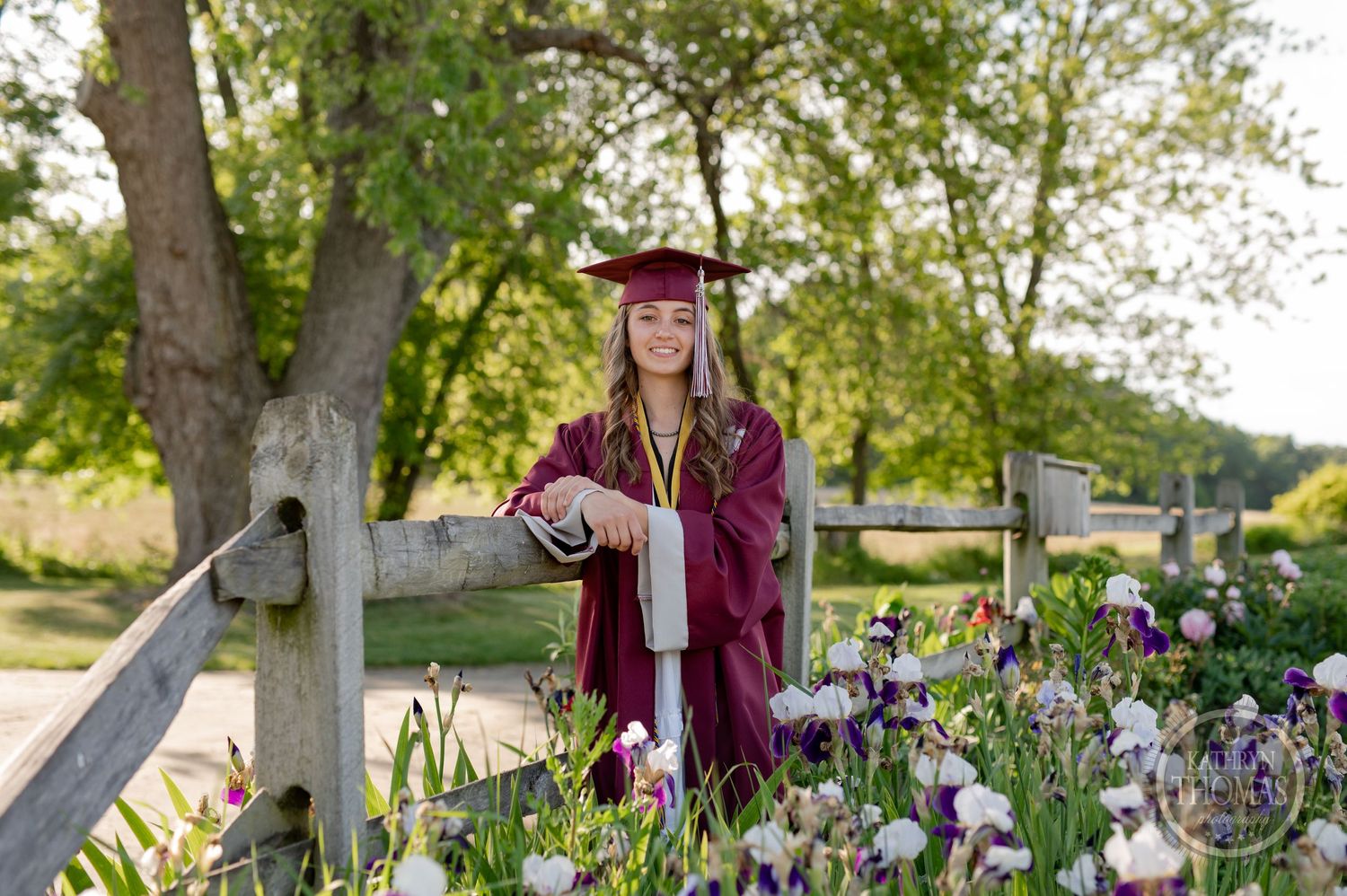 Graduate in maroon regalia stands among blooming iris flowers in garden setting.