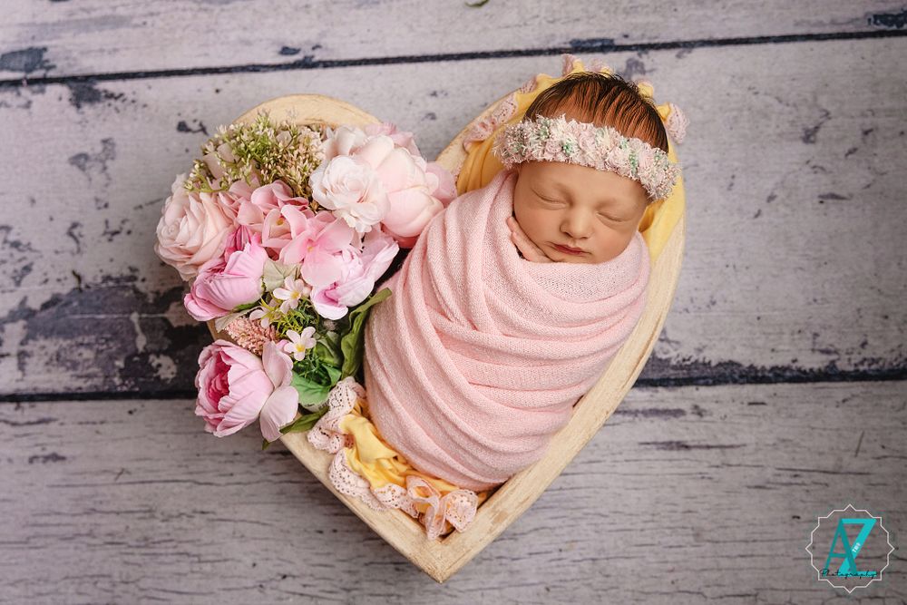 Newborn in pink wrap sleeping in heart-shaped basket with pink roses on rustic wood background.
