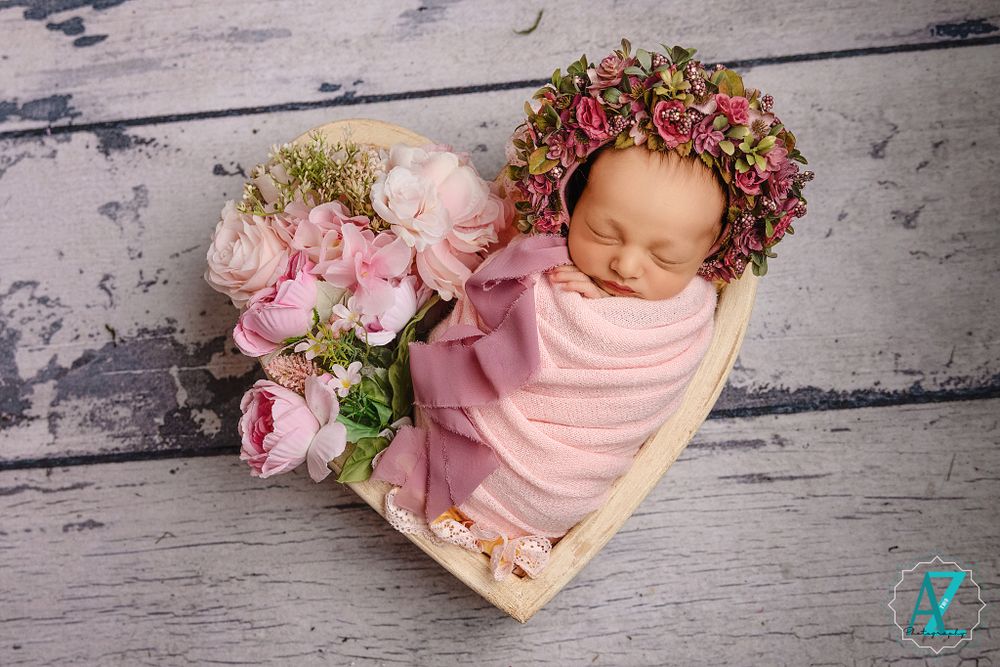 Pink-wrapped newborn rests in wooden heart bowl with pink roses and floral crown on rustic background.