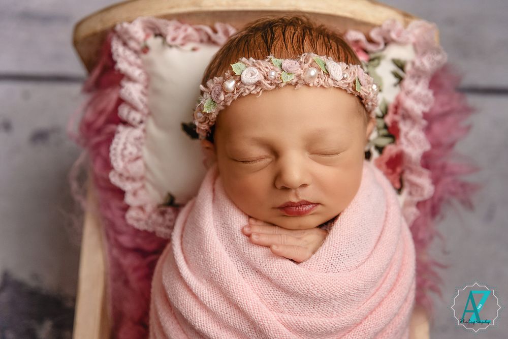 Sleeping newborn in pink wrap wearing delicate floral headpiece on lace backdrop.