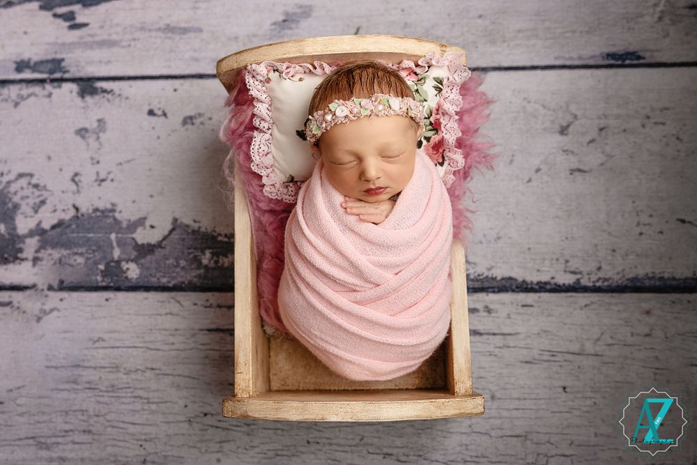 Peaceful newborn swaddled in pink fabric with floral headband resting in wooden prop.