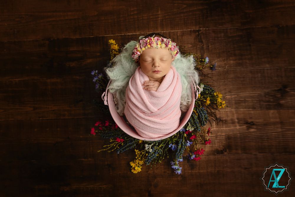 Overhead view of newborn in pink wrap surrounded by colorful wildflowers on dark wooden surface.