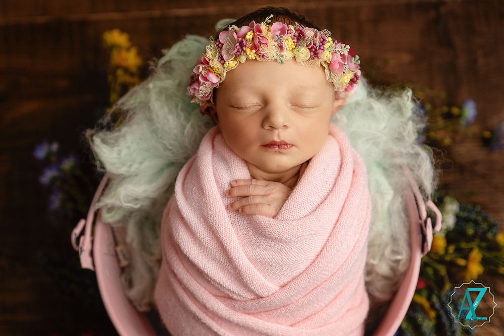 Close-up of sleeping baby wrapped in pink fabric with floral crown and soft white bedding.