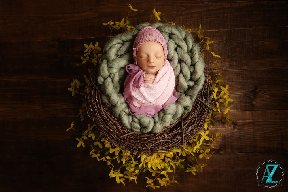 Sleeping newborn in pink wrap nestled in woven nest surrounded by yellow flowers.