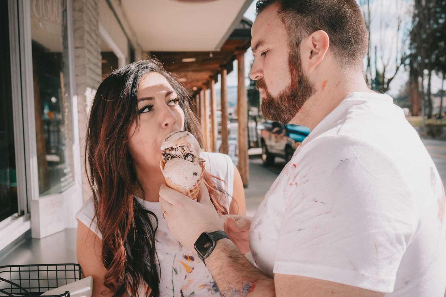 A couple is sharing an ice cream cone he is feeding her they are covered in paint. They are at Scoop There It Is in Orting