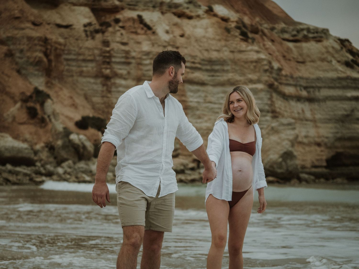 Pregnant woman with partner walking along beach whilst smiling at each other