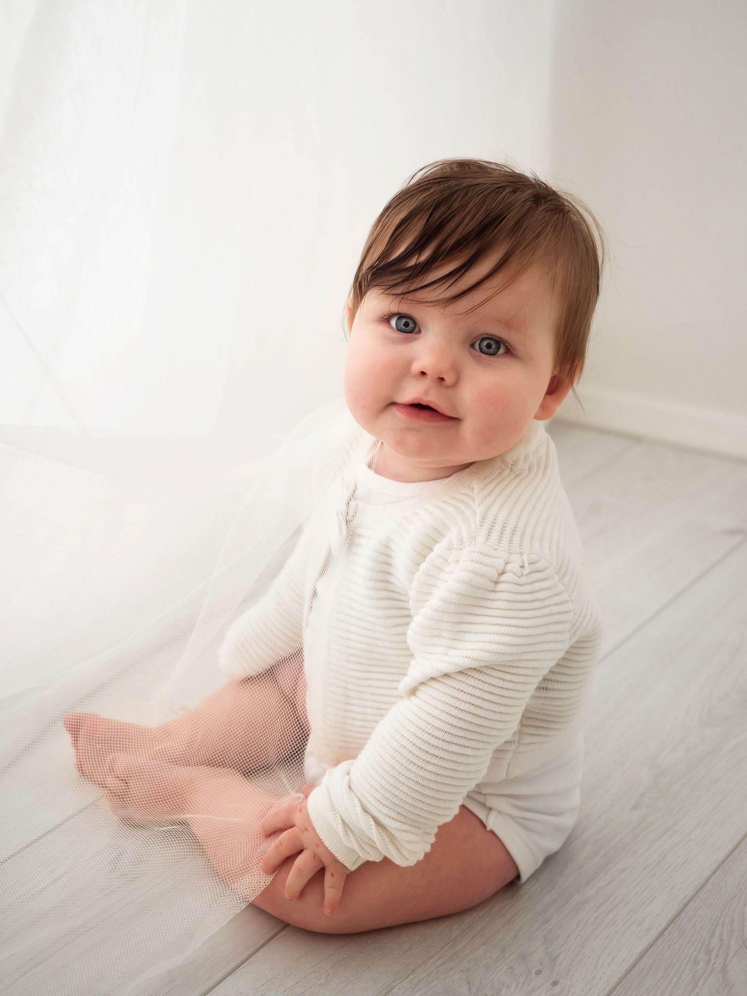 Baby girl in white jumper sits by white curtain during Didcot photoshoot