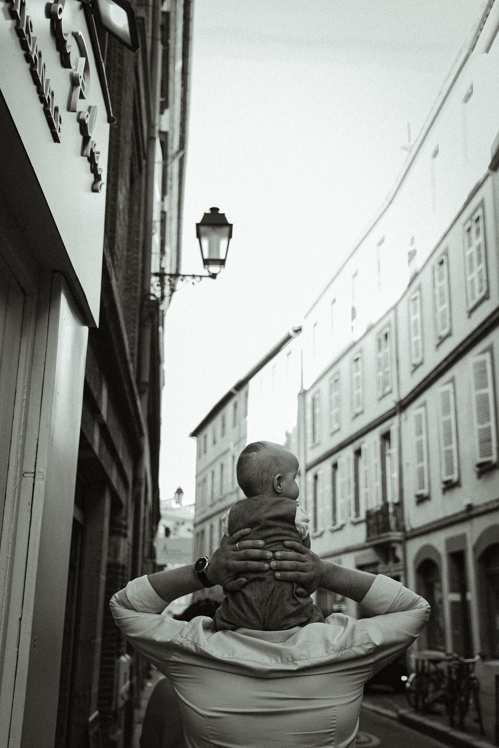 Black and white photo of baby riding on shoulders through city street with vintage lamp post.