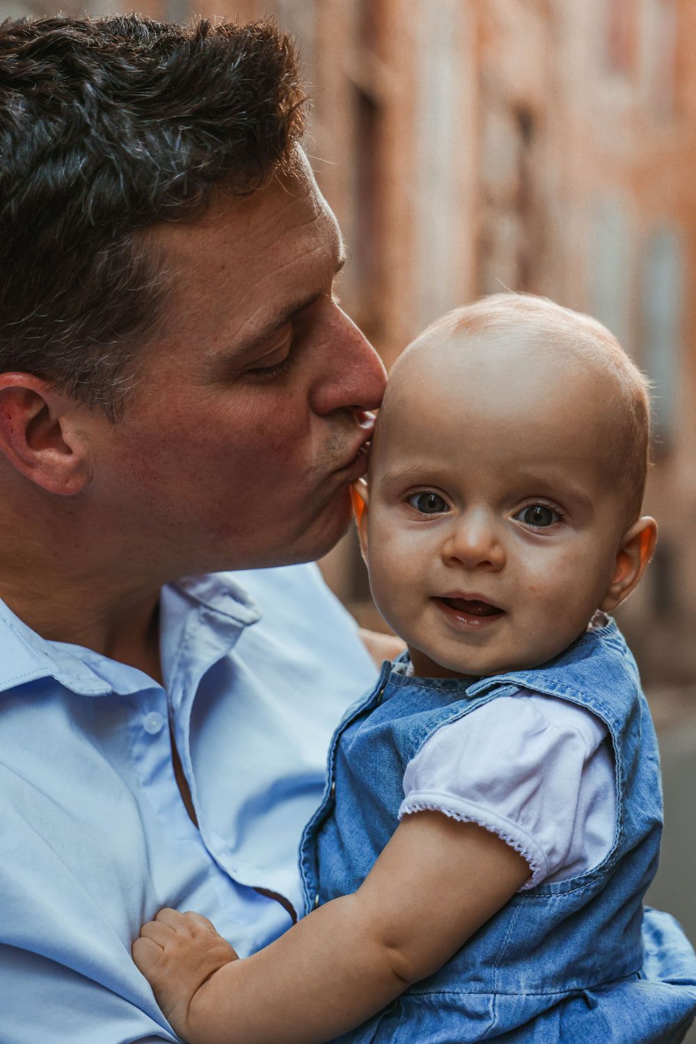 A tender moment captured as a baby in blue clothing receives a gentle kiss on their head.
