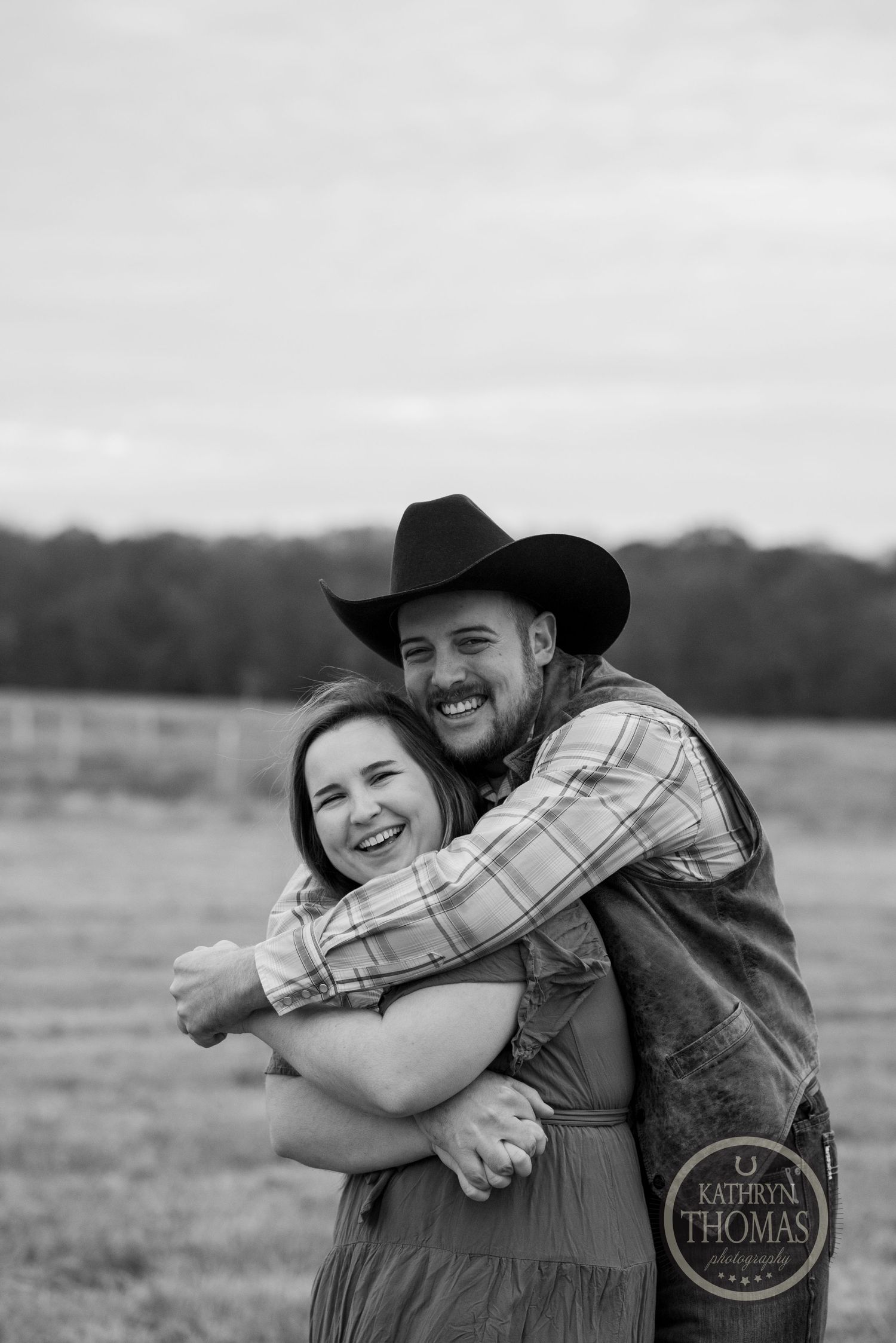 A joyful black and white photo of a couple laughing and embracing in a country field.