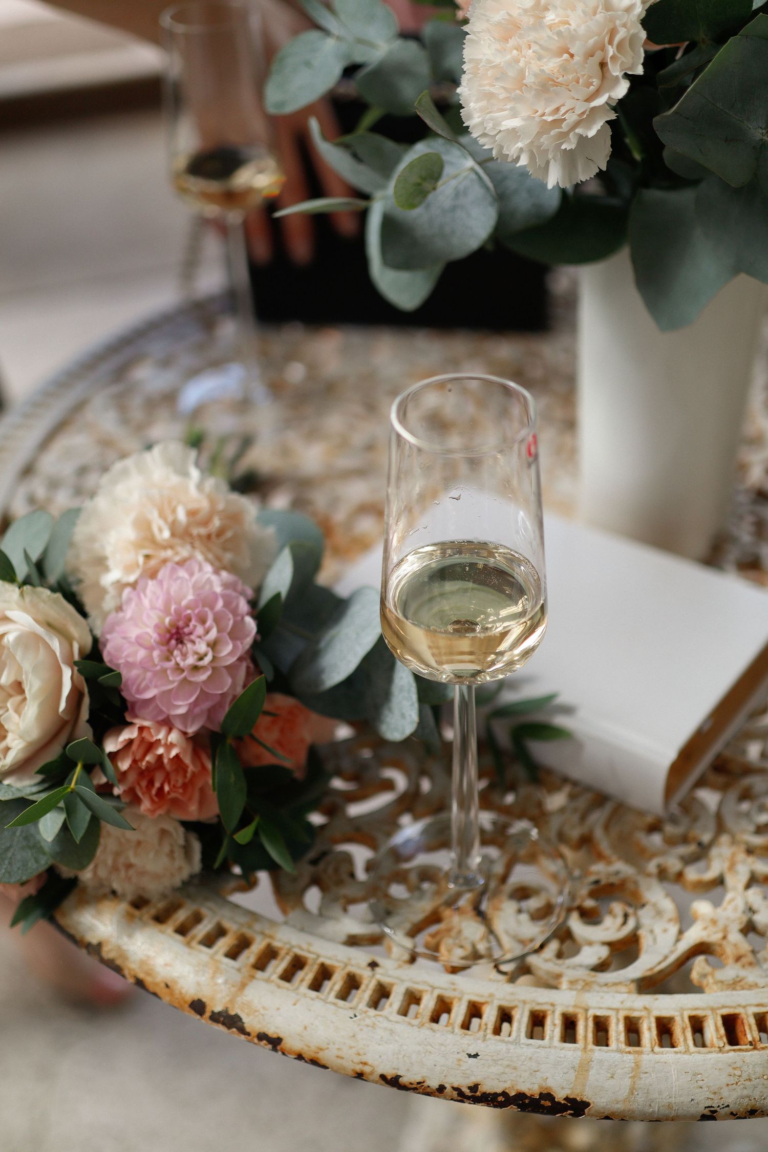 Elegant floral arrangement with white and pink carnations beside a glass of white wine on an ornate tray.