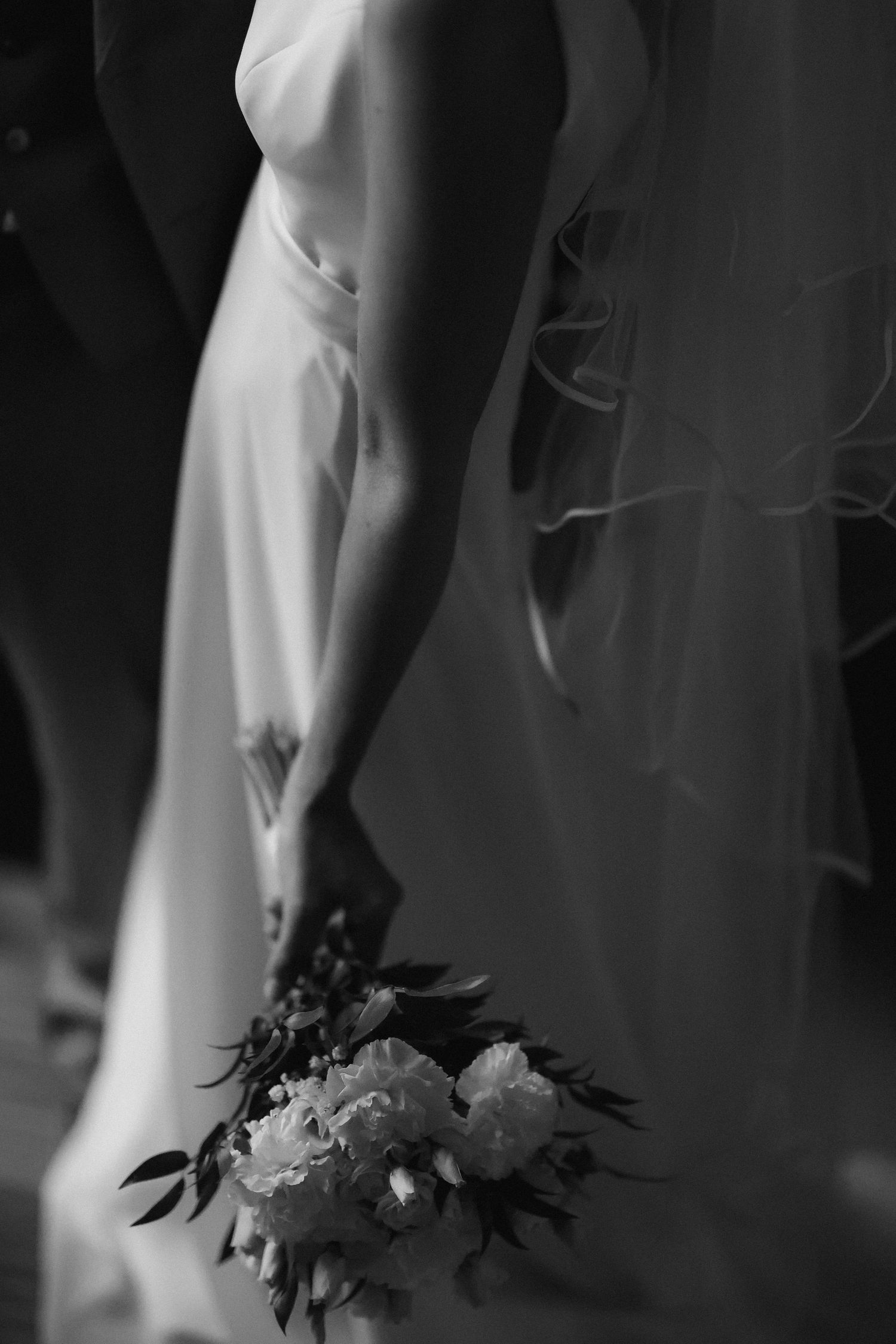 Black and white artistic photo of a bride holding a bouquet, showing the elegant drape of her dress.