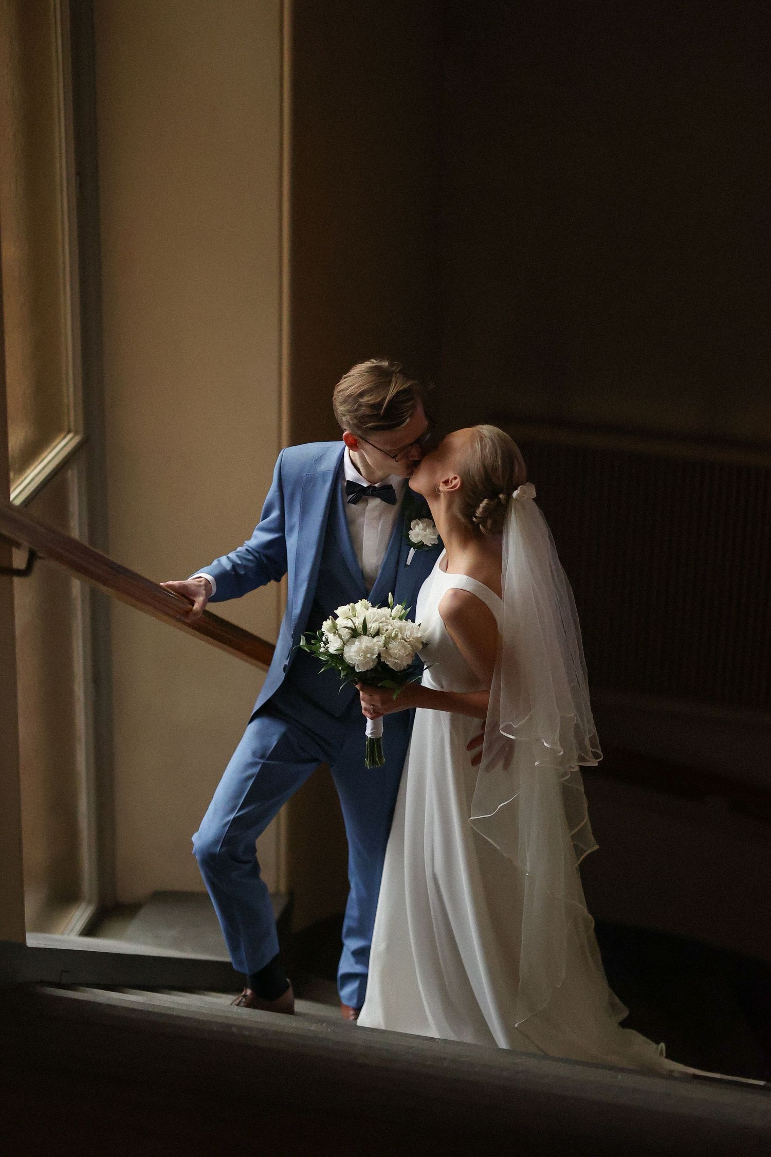 A couple in wedding attire shares a romantic moment on a staircase in dramatic window light.