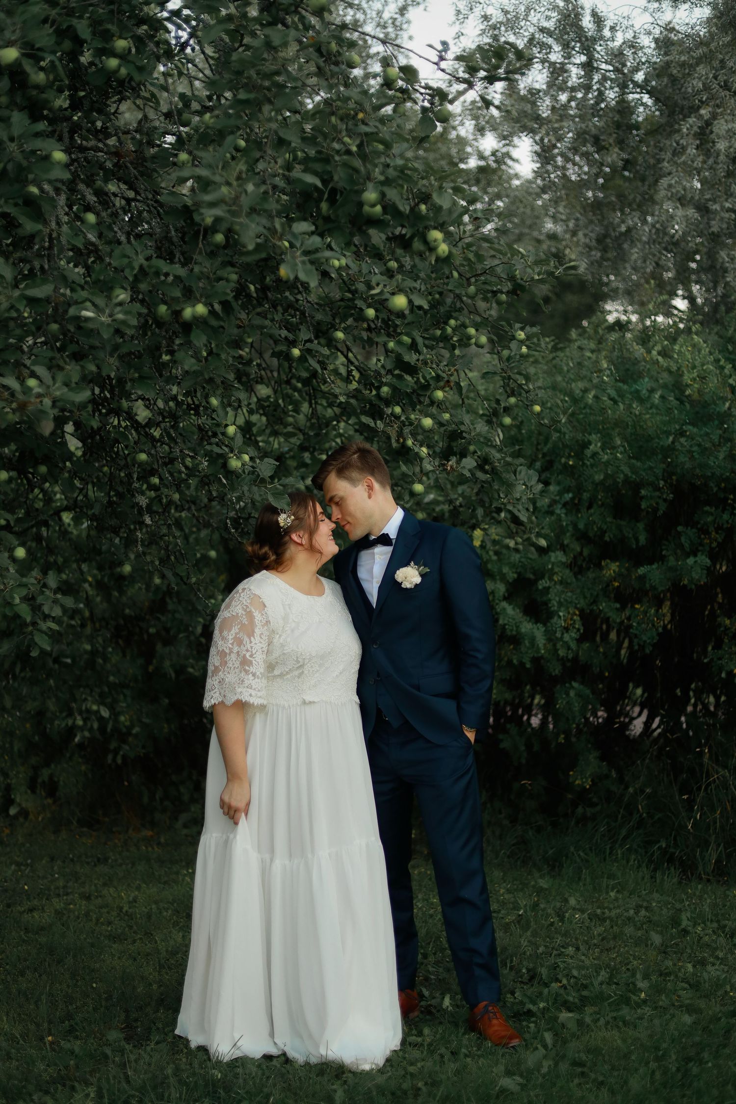A couple poses together in formal wedding attire against a lush green garden backdrop.