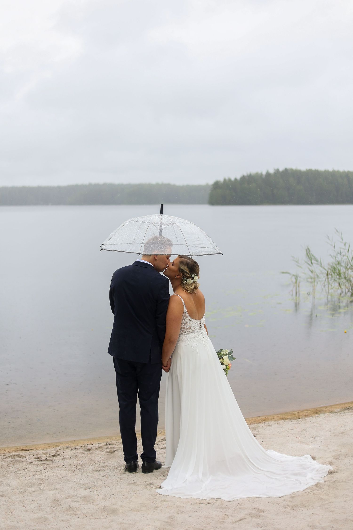 A couple shares an intimate moment under a clear umbrella by a misty lake.