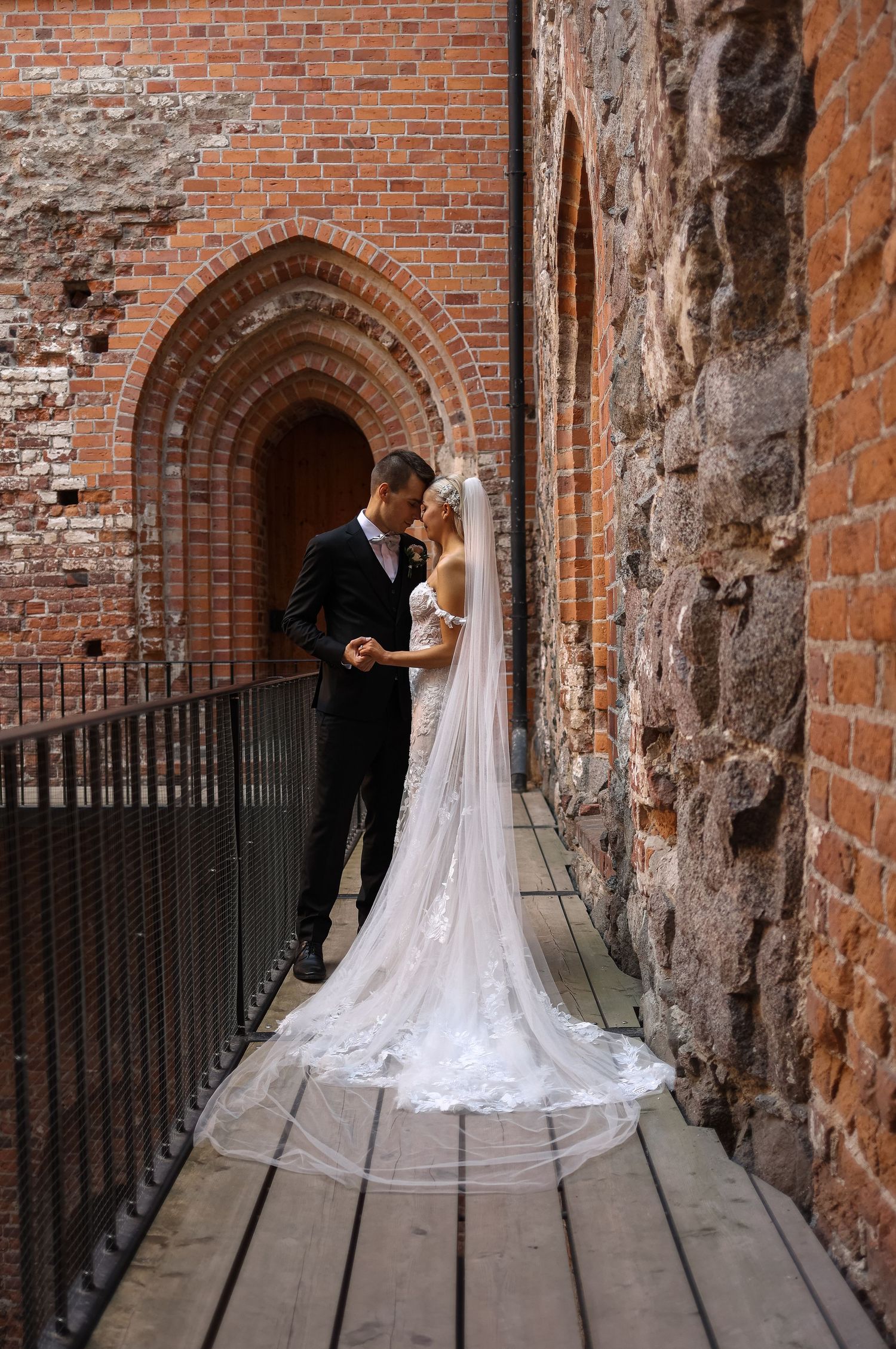 A wedding couple embraces in a historic brick archway with a flowing white veil trailing behind.