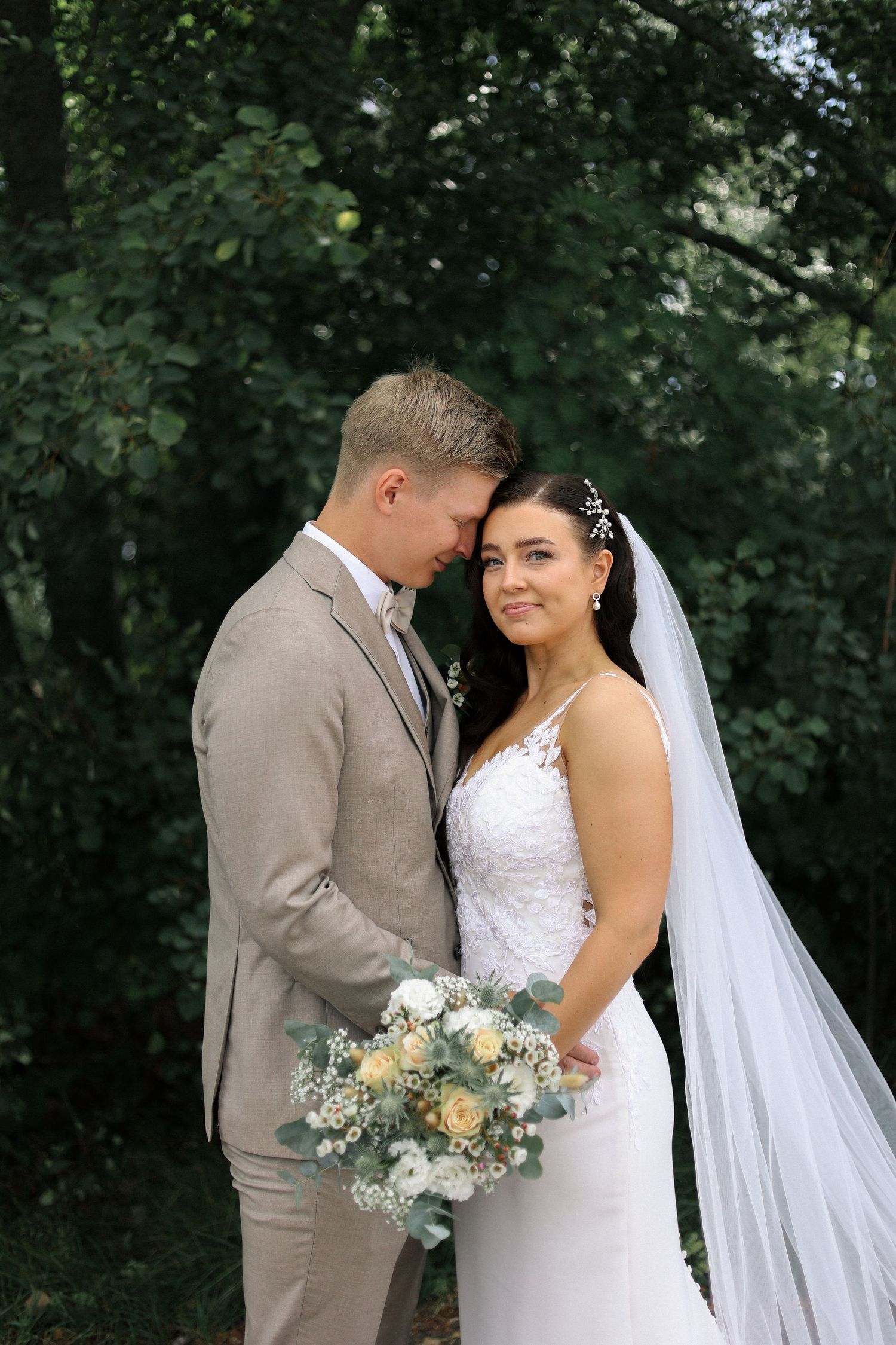 A romantic wedding photo of a couple in a garden setting with a soft beige suit and white lace dress.