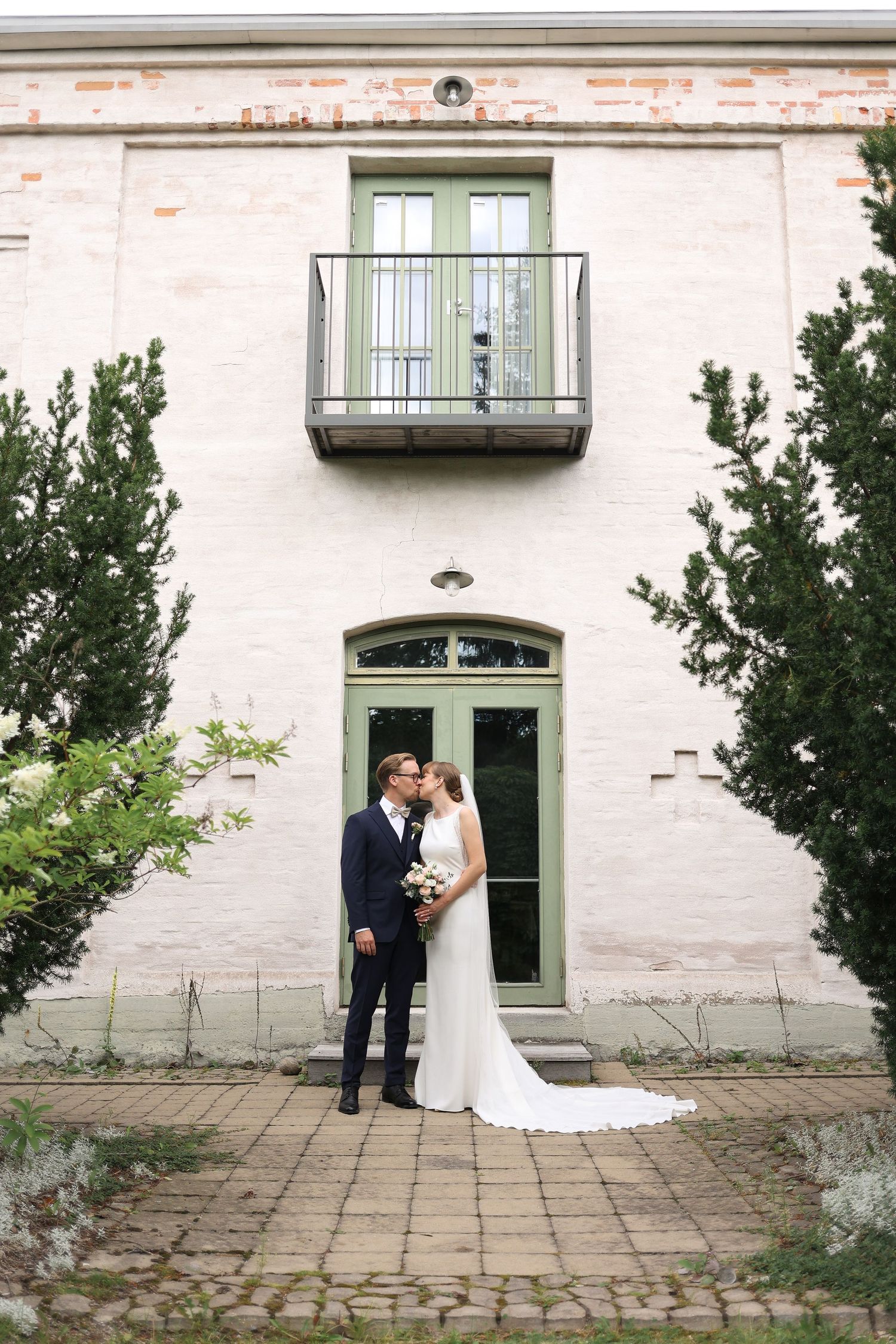 A wedding couple poses in front of a vintage building with green doors and balcony.