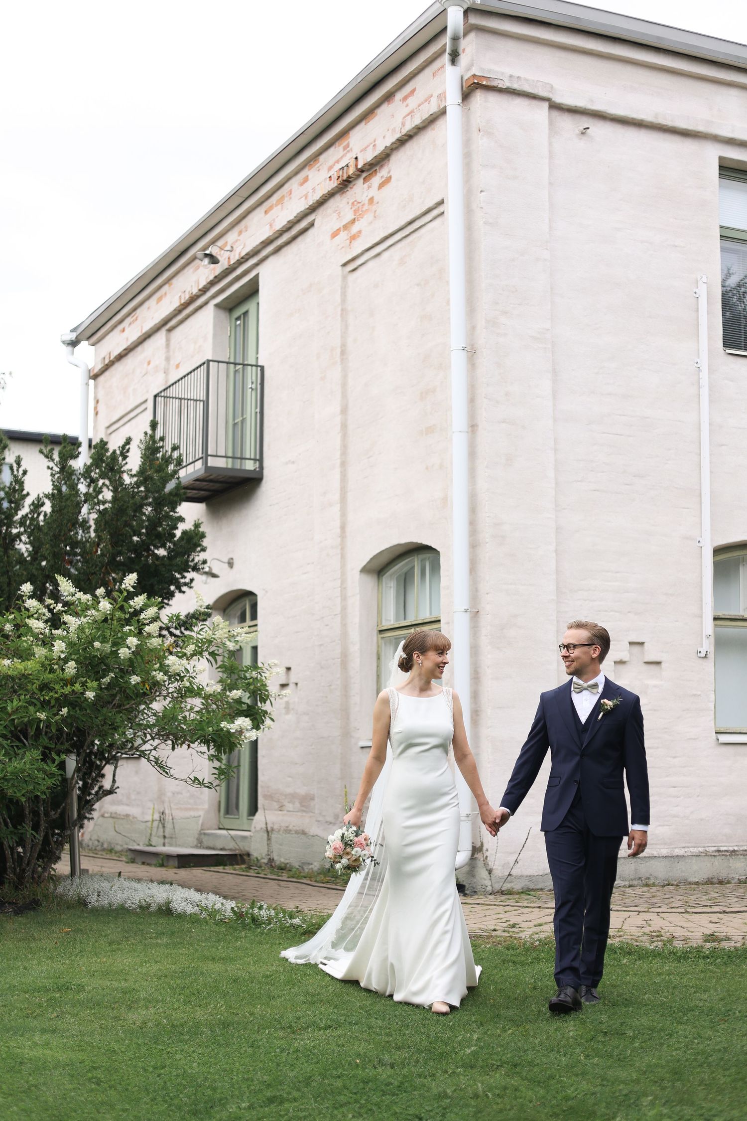 A newlywed couple walking hand in hand on grass in front of a historic building.