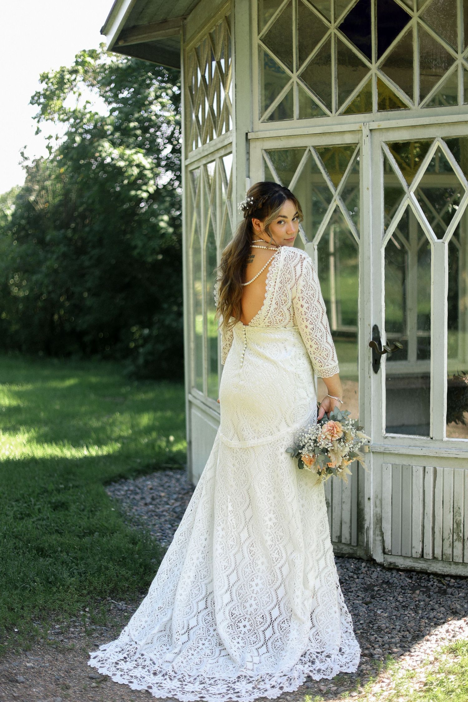 Bride in elegant lace wedding gown poses by vintage gazebo in garden setting.