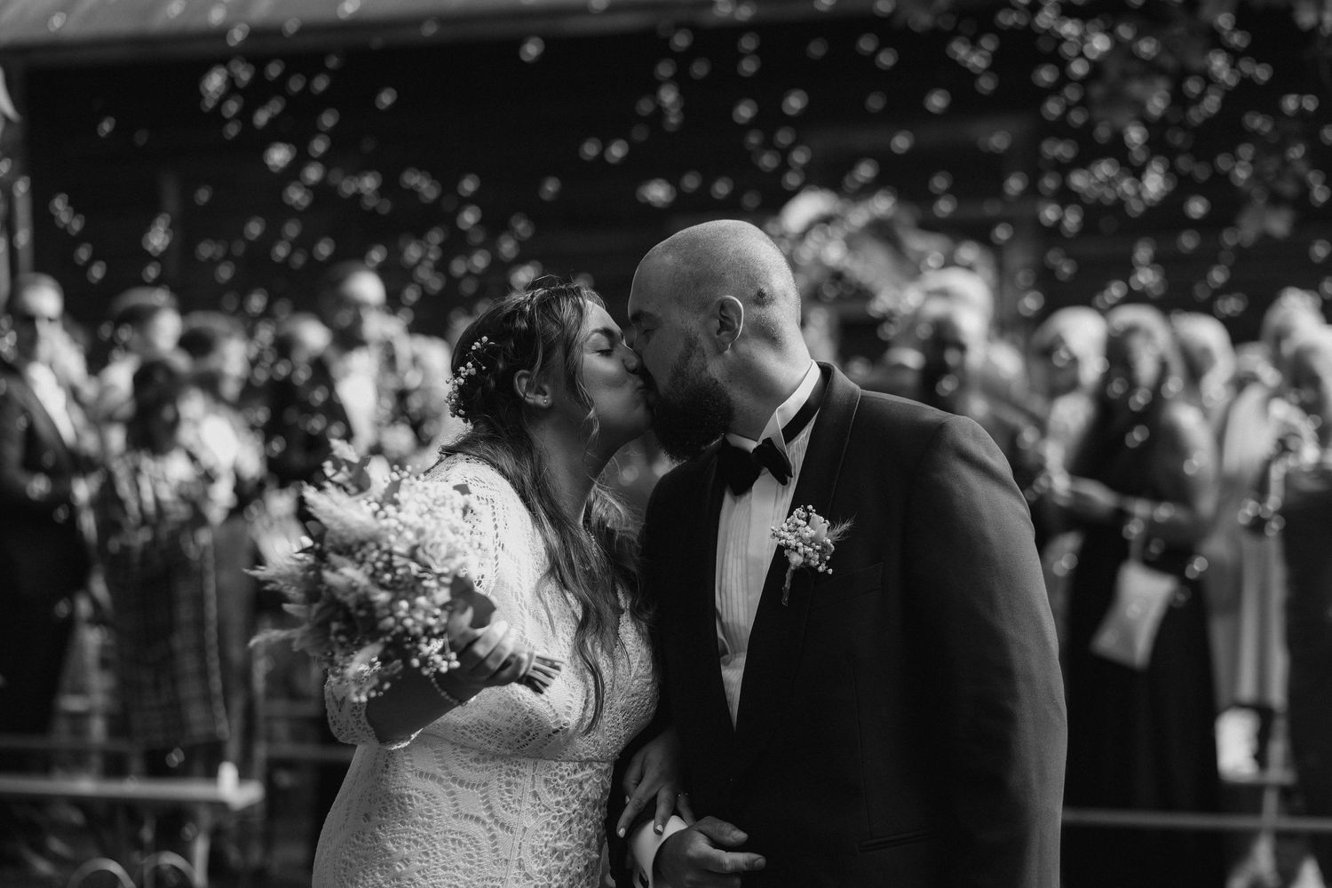 Black and white wedding photo capturing tender moment between newlyweds with crowd in background.