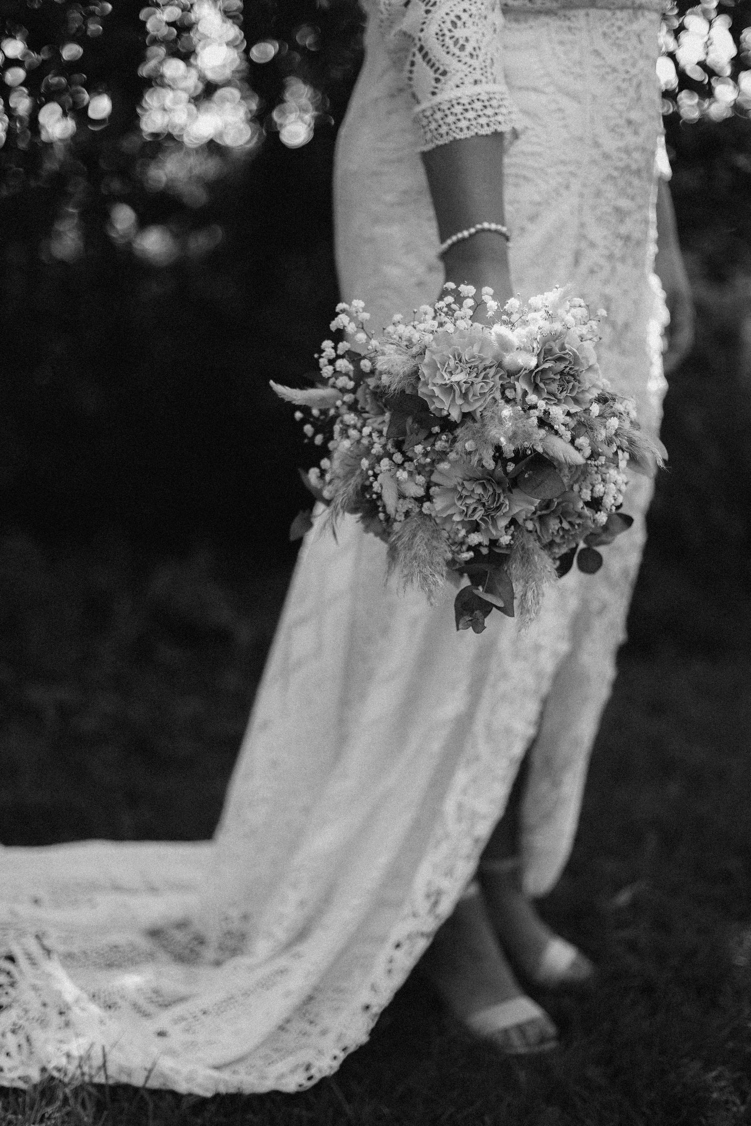 Black and white detail shot of bride holding delicate floral bouquet with lace dress train.