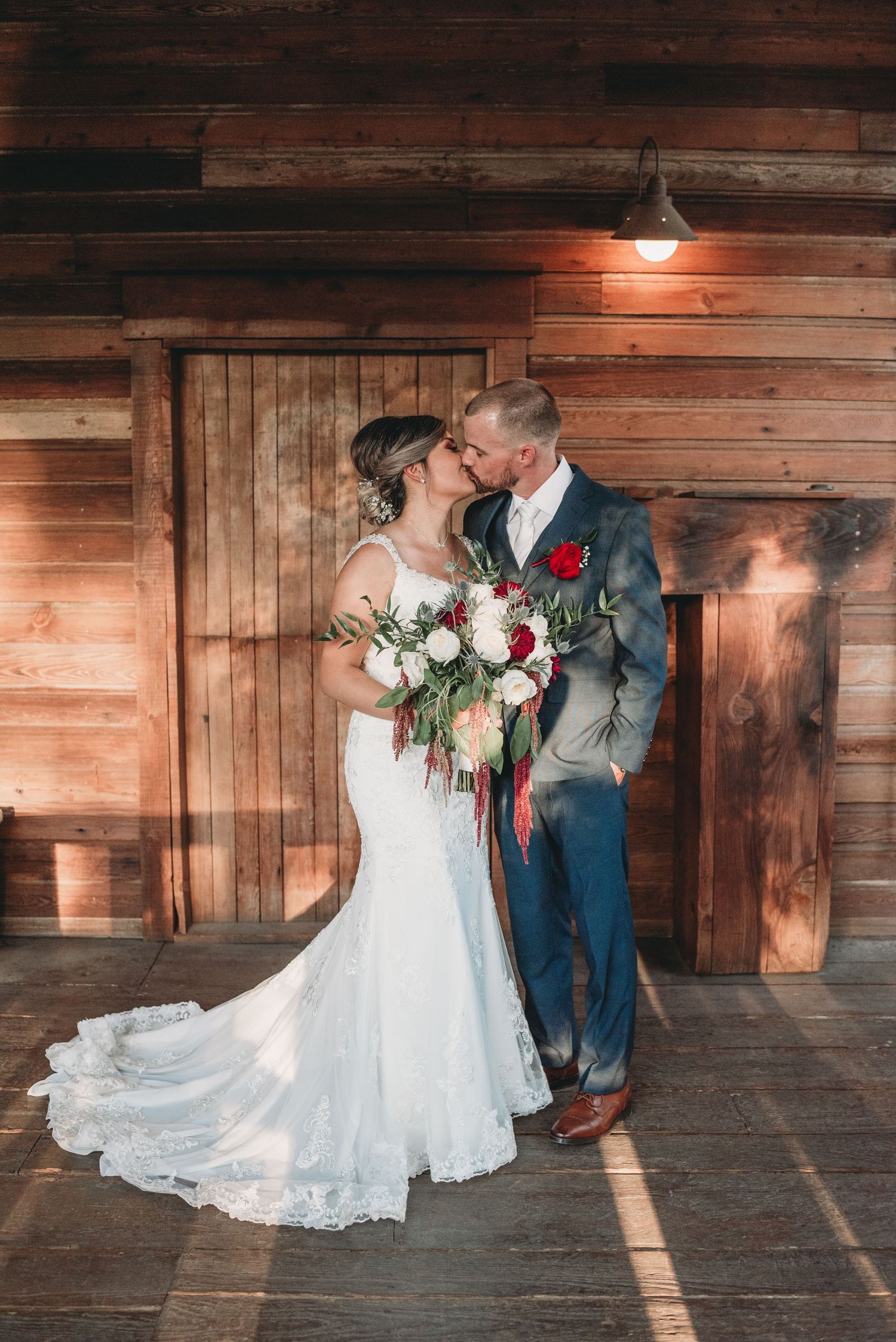 Romantic wedding couple embraces in rustic wooden barn with dramatic lighting and white lace gown.