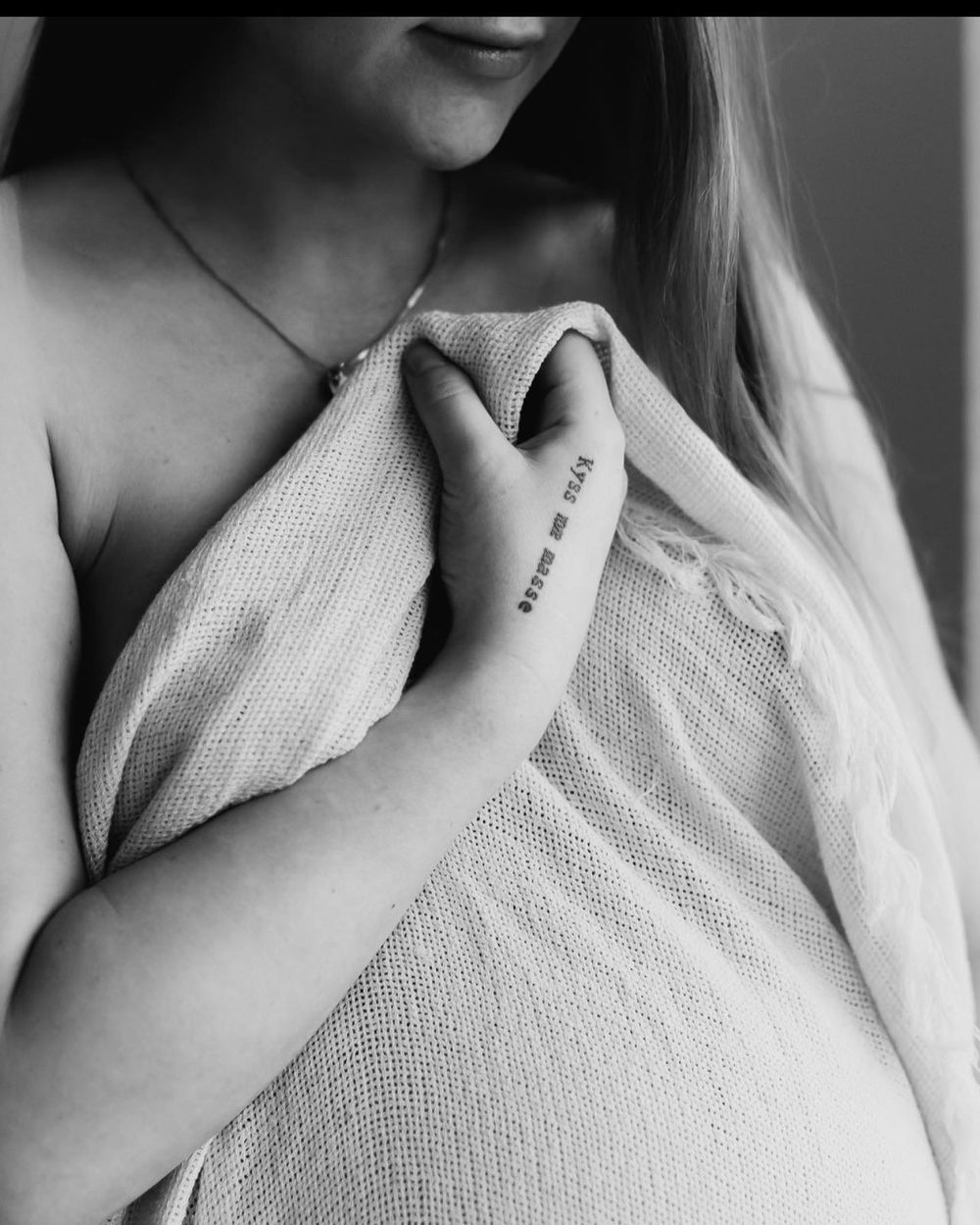 Svart-hvitt-bilde som viser en hånd med liten tatovering og en gravidmage under et lett teppe