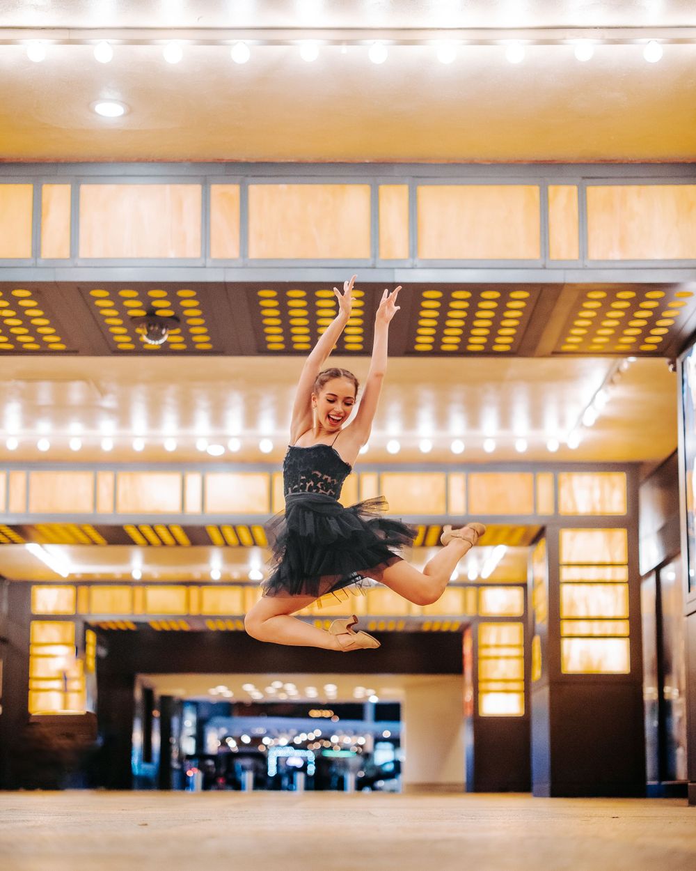 Performer in black tutu leaps gracefully beneath illuminated theater marquee.