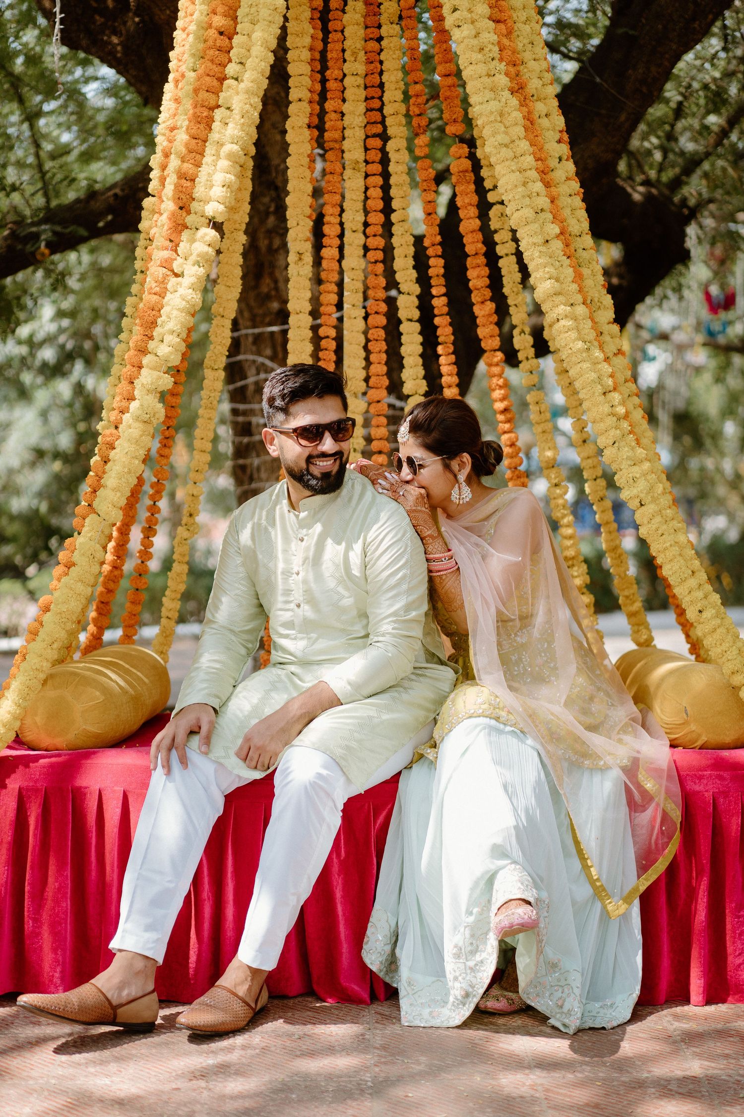 Couple sits beneath orange marigold garland decorations on red-draped seating during traditional ceremony.