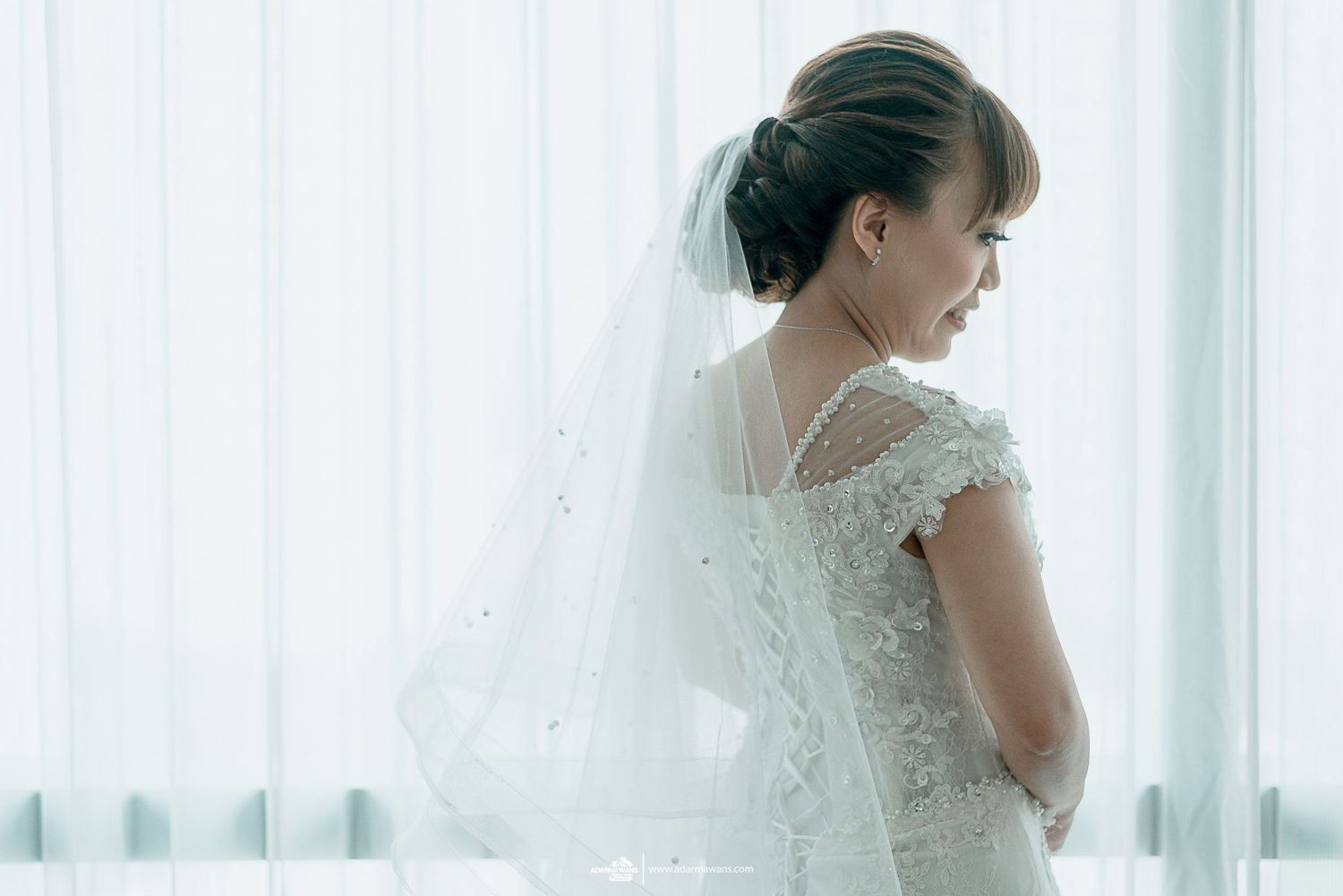 A bride in a detailed lace dress stands thoughtfully by a window with flowing white curtains.