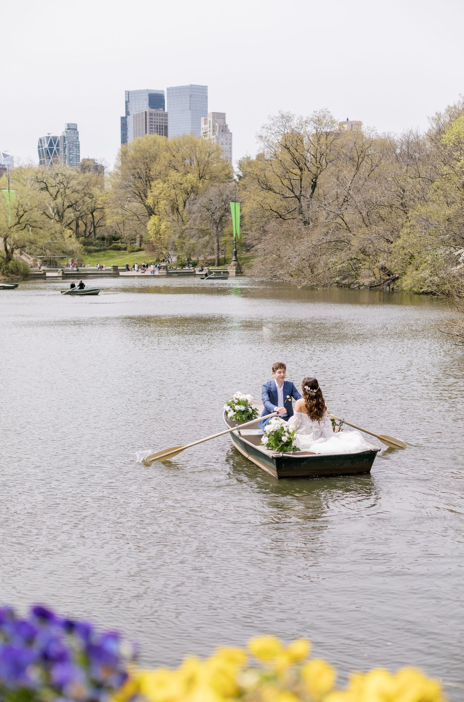 Newlyweds rowing a boat on a Central Park lake with city skyline in background.