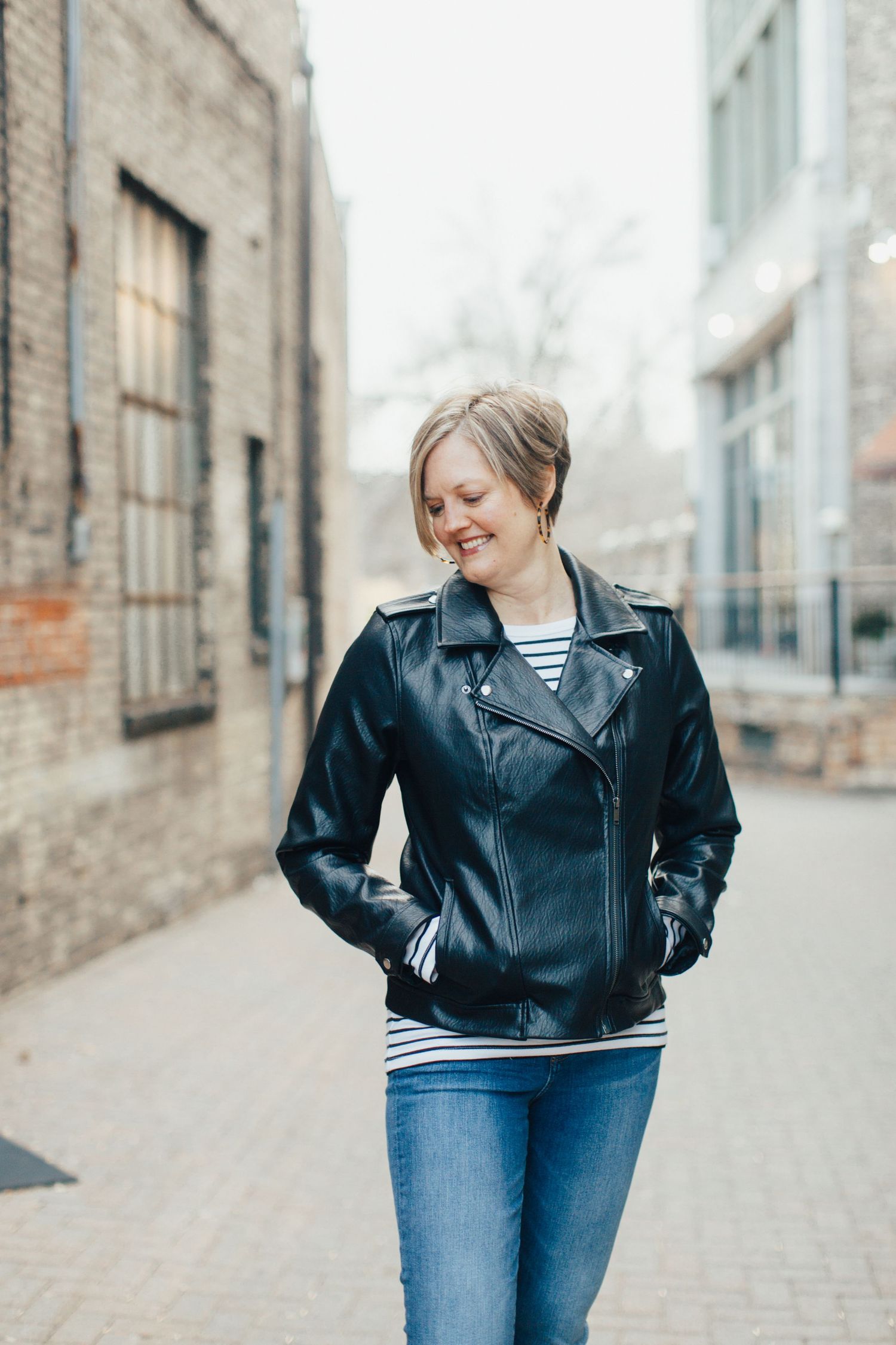 Model wearing classic black leather jacket and blue jeans poses casually against urban brick building backdrop.