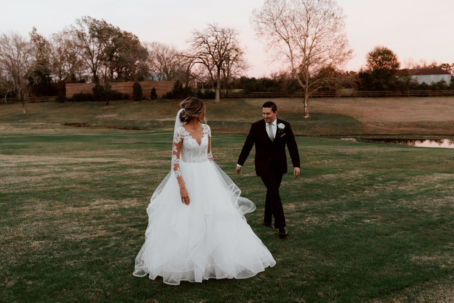 A couple in wedding attire walks hand in hand across a lush green field at sunset with bare trees in the background.