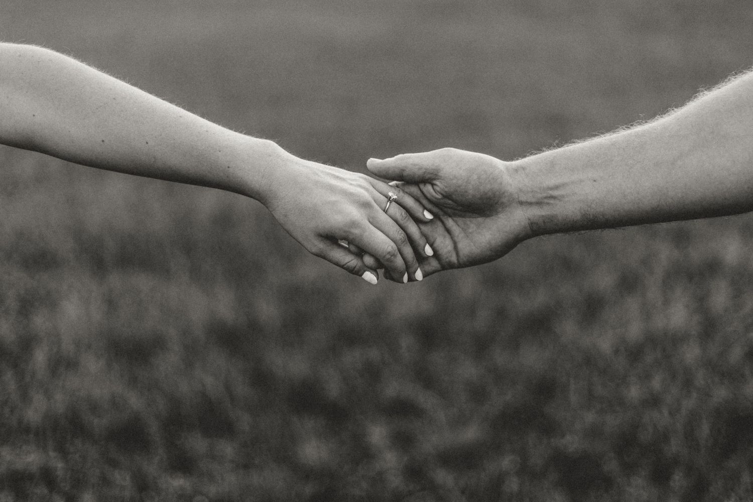 A black and white close-up of two hands touching intimately in a grassy field.
