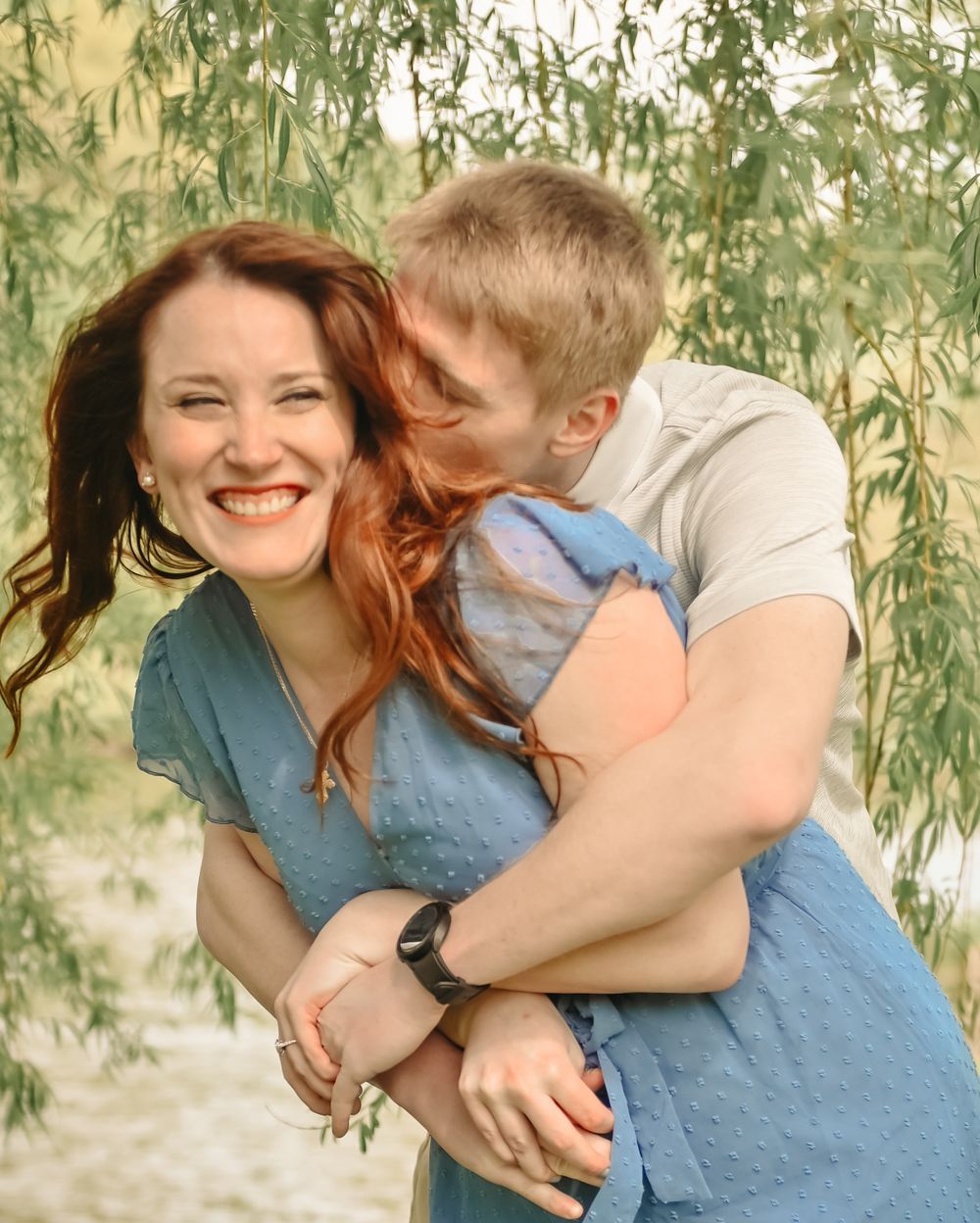 A joyful romantic embrace under willow branches with a couple wearing blue and white casual attire.