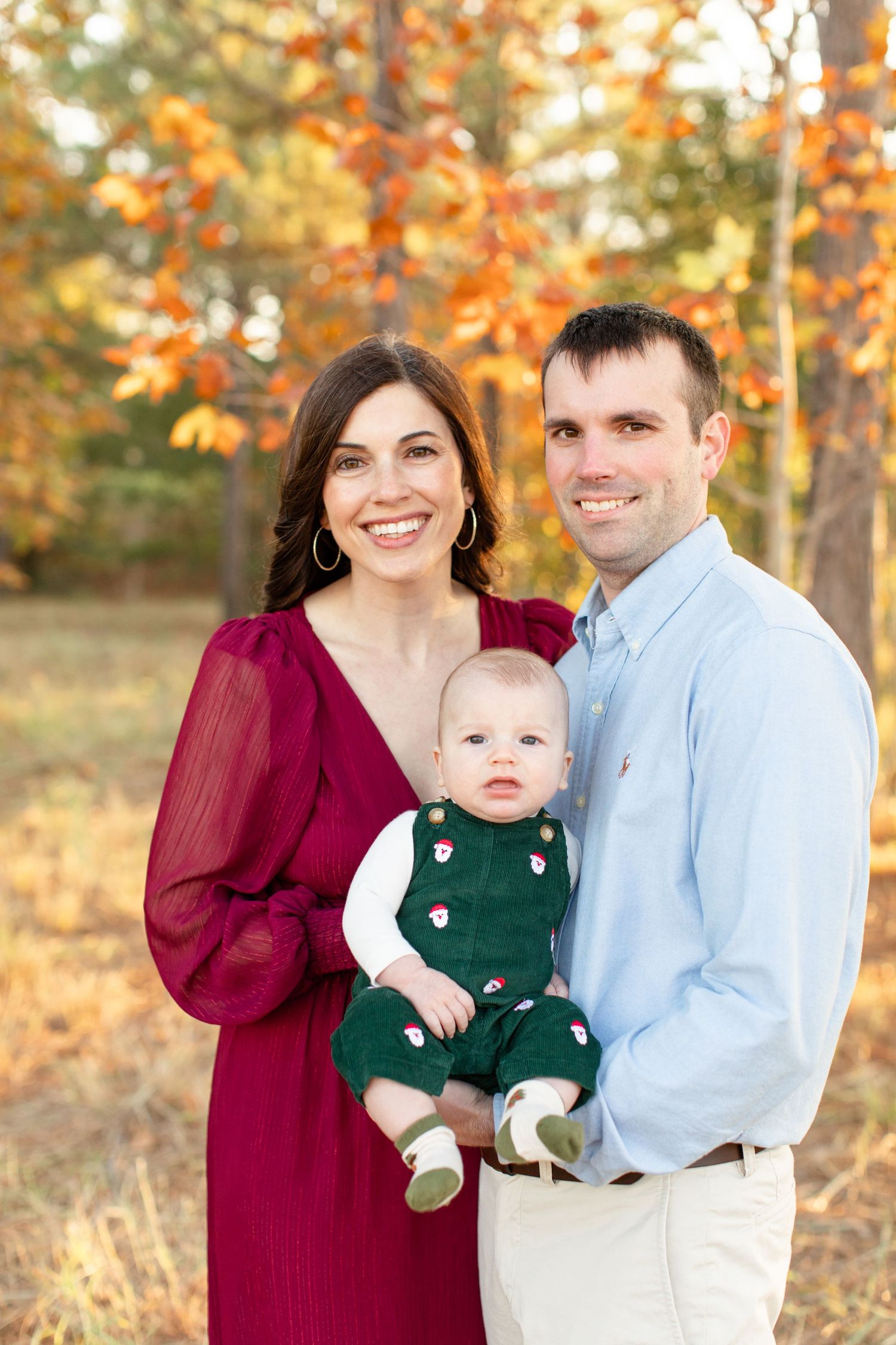 A family portrait in front of autumn trees with parents in burgundy and light blue holding a baby in green overalls.