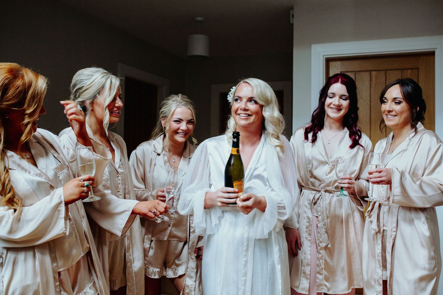 Group in white silk robes celebrating with champagne before a wedding ceremony.