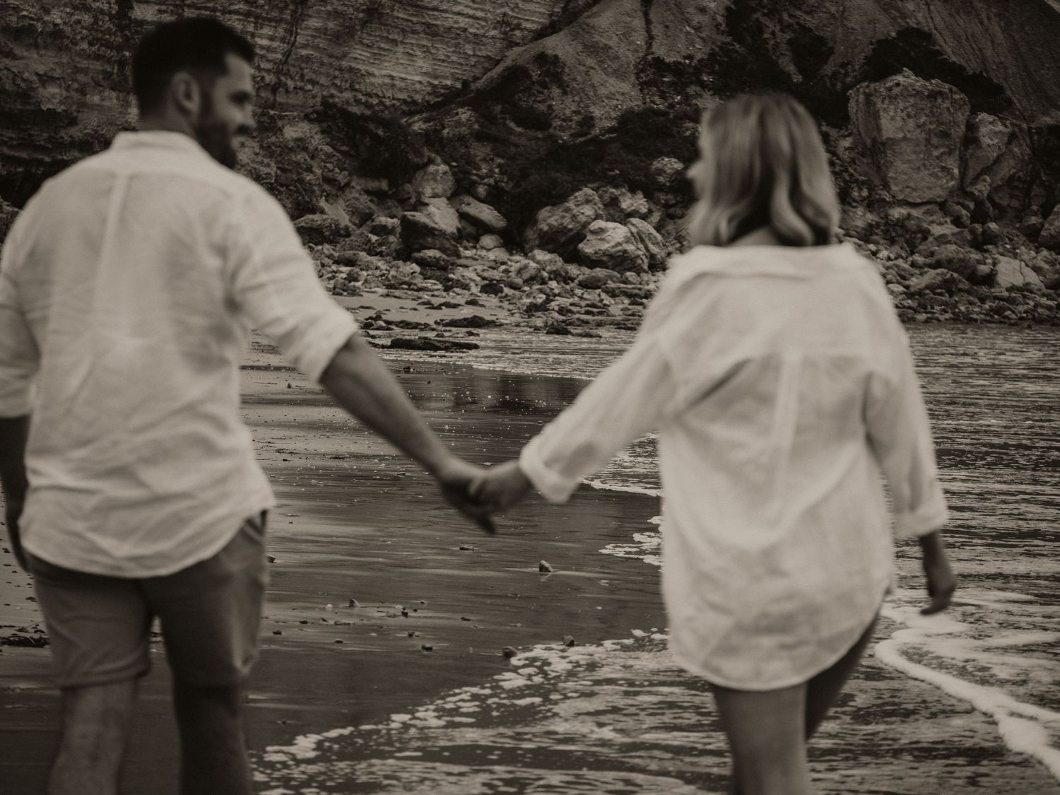 A black and white beach scene showing a couple holding hands near rocky cliffs.