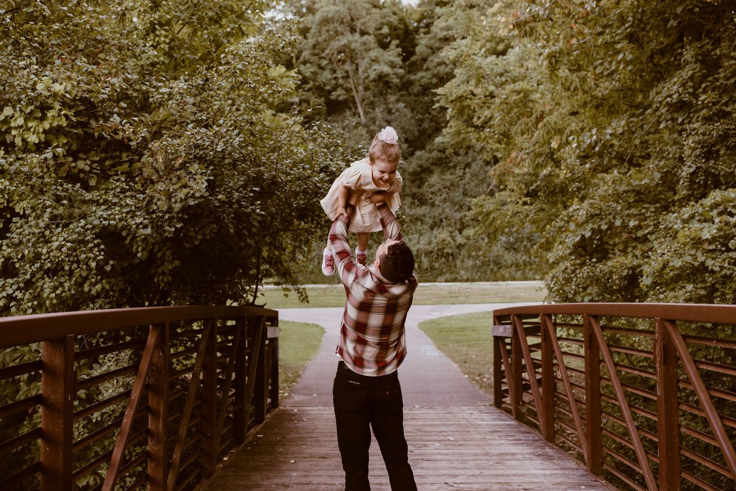 A parent lifts a child high in the air while standing on a wooden bridge surrounded by trees.