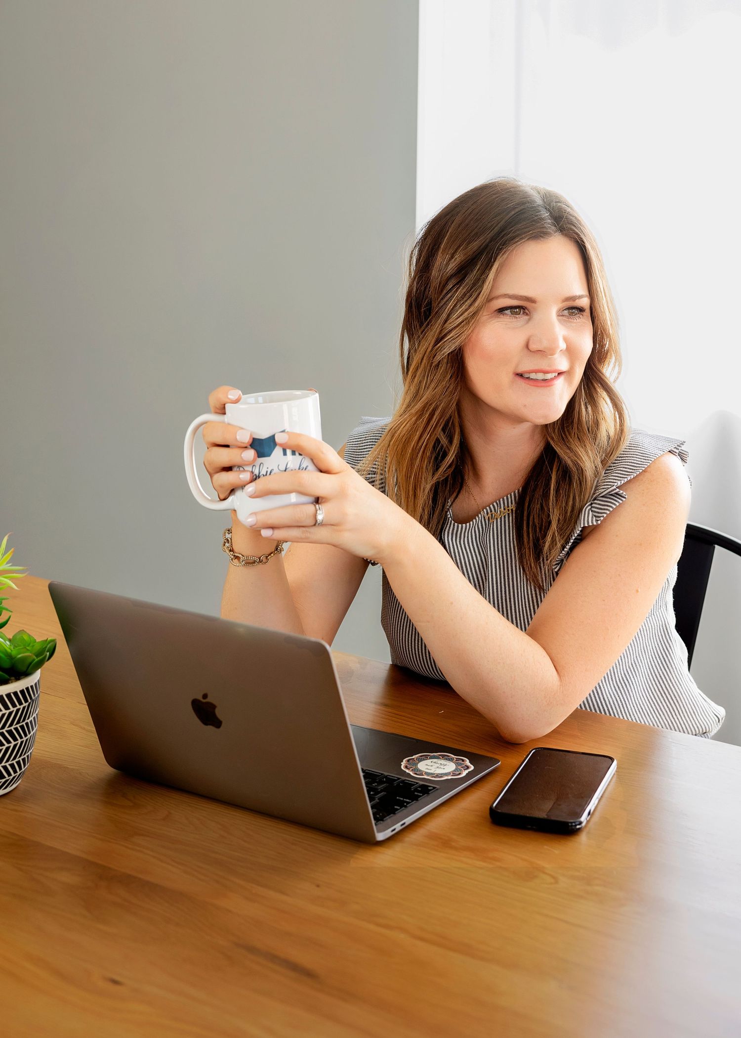 Person working at wooden desk with MacBook and coffee mug in bright workspace.