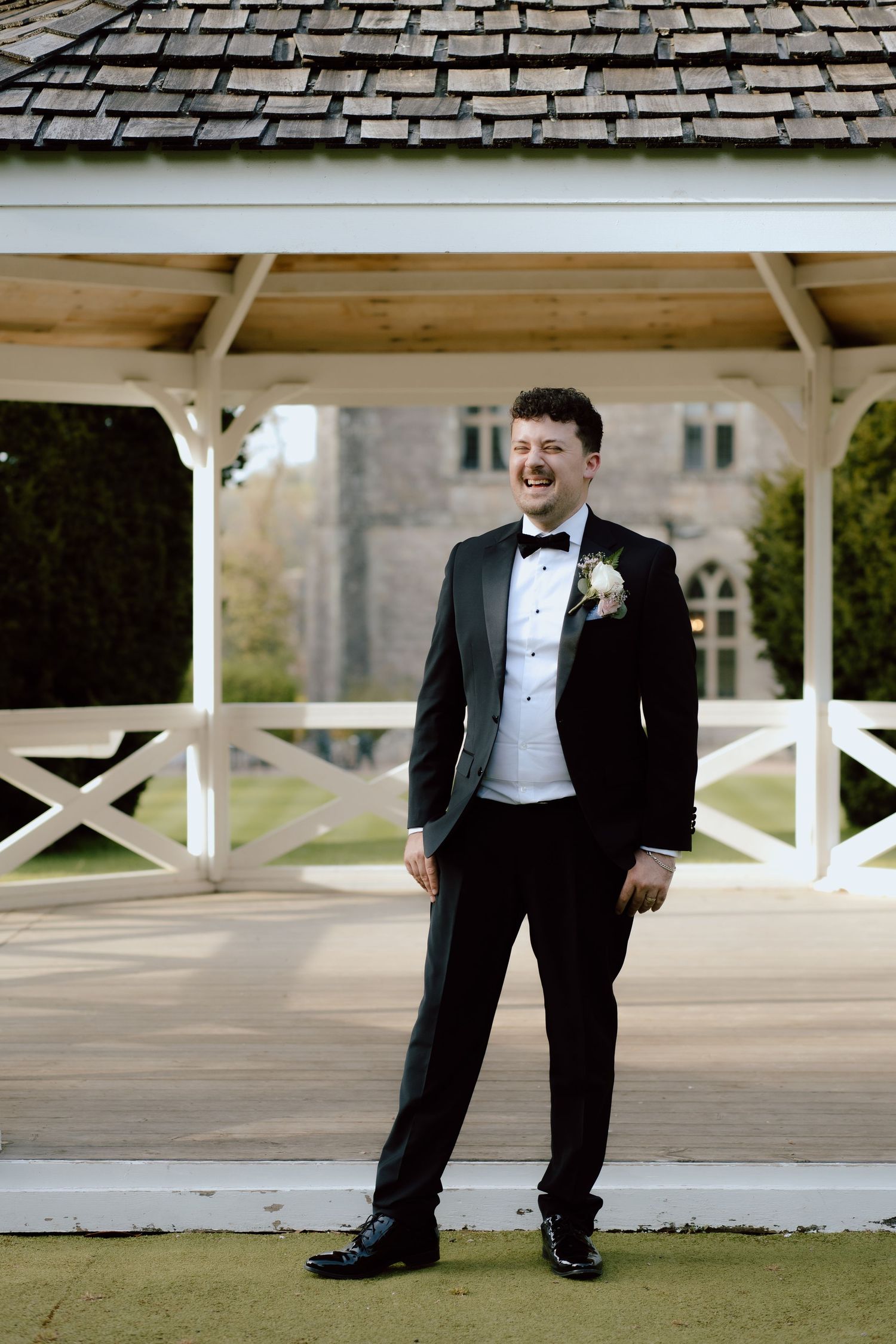 A groom in classic black tuxedo poses under a white gazebo on wedding day.