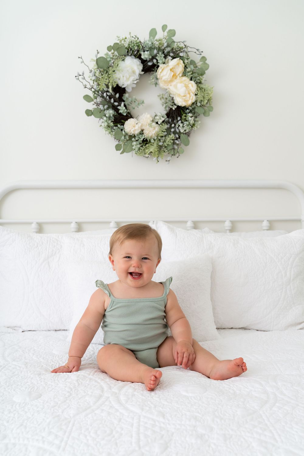 Joyful baby girl in sage green romper sits on a white bed below a floral eucalyptus wreath.